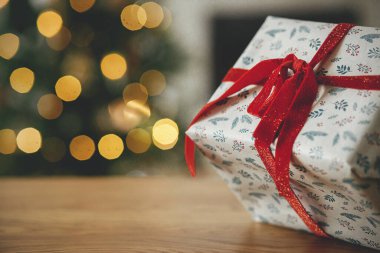 Mutlu Noeller! Şık bir Noel hediyesi, arka plandaki altın ışıklara yakın çekim. Ahşap masada kırmızı kurdeleyle sarılı modern hediye, yazı için yer. Tatil afişi