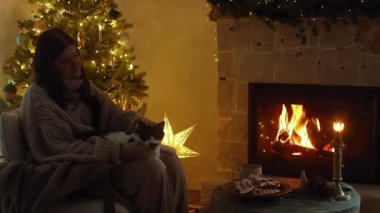 Kucağındaki sevimli kediyi okşayan, Noel arifesinde yanan şöminede gevşeyen hoş kazaklı bir kadın. Atmosferik Noel arifesi görüntüleri. Evcil hayvan ve kış tatili