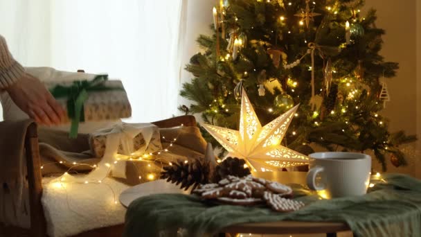圣诞快乐 手穿舒适的毛衣 把时尚包装的礼物放在现代的椅子上 在喜庆的圣诞树上 舒适的房间里点着发光的灯 大气圣诞前夕的镜头 — 图库视频影像