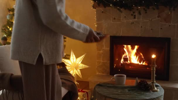 お祝いの暖炉とクリスマスツリーライトに対して おいしいジンジャーブレッドクッキーとティーカップをテーブルに置く居心地の良いセーターの女性 大気クリスマスイブ映像 — ストック動画