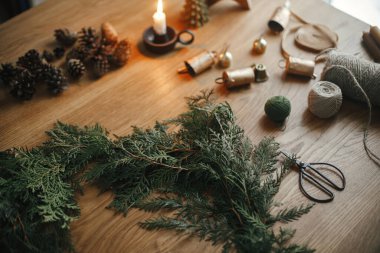 Noel kırsal çelengi yapıyorum. Çember, sedir dalları, kozalaklar, mum, sicim, çanlar, makas ahşap masa üzerinde karamsar festival odasında. Kış tatili hazırlıkları, atmosferik zaman