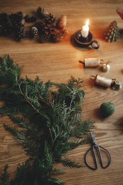 Firavun dalları, vintage çanlar, kozalaklar, sicim, mum ve makas, ahşap masada, yakın çekim. Noel kırsal çelengi yapıyorum. Akşam atmosferik odasında kış tatili hazırlıkları