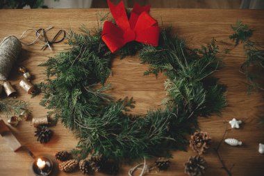 Kırmızı yay, sedir dalları, vintage çanlar, çam kozalakları, sicim, ahşap masa üzerinde makas ile süslü Noel rustik çelengi. Mutlu noeller. Atmosferik odada kış tatili hazırlıkları