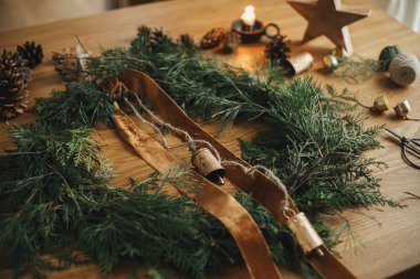 Sedir dalları, kurdele, eski çanlar, çam kozalakları, sicim, ahşap masa üzerinde makas olan şık bir Noel rustik çelengi. Mutlu noeller. Atmosferik odada kış tatili hazırlıkları