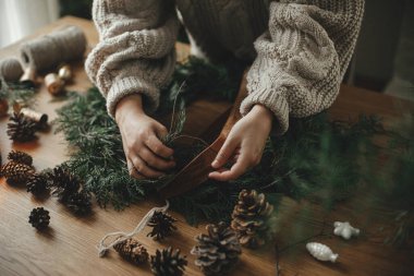 Noel kırsal çelengi hazırlıyorum. Kurdele tutan eller, sedir dalları, çam kozalakları ile ahşap masa üzerinde çelenk yapmak, atmosferik festival odasında çanlar. Kış tatili hazırlıkları