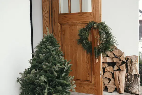在木制前门和冷杉树上的黄铜把手上挂着老式铃铛和彩带的别致的圣诞乡村花环 冬季农村假日的现代生态农舍外部装饰 — 图库照片