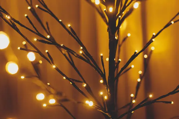 Stylish minimalist glowing tree in evening cozy room. Scandinavian christmas decor, minimal modern black tree branches golden lights bokeh, copy space. Happy Holidays! Festive illumination