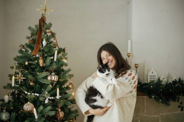 Şenlik odasında süslü Noel ağacının arka planında sevimli kediyi kucaklayan rahat kazaklı mutlu kadın. Hayvan ve kış tatillerinin sahibi. Mutlu Noeller.!