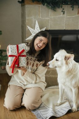 Hoş süveterli bir kadın Noel hediyesini şık bir Noel ağacı ve şöminesinin arkasında beyaz sevimli bir köpekle açıyor. Kış tatili Kutlama odasında evcil hayvan sahibi. Mutlu Noeller.!