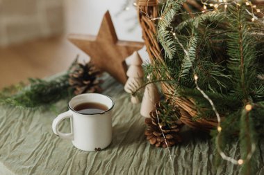 İskandinav kızlık zarı. Şık bir fincan sıcak çay, sepetteki köknar dalları, tahta ağaçlar ve yıldız, kozalaklar ve masa ışıkları. Modern Noel rustik çevre dostu dekor ve kupa modeli