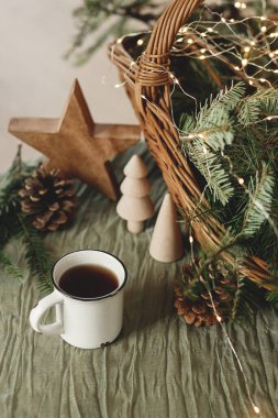 Şık bir fincan sıcak çay, sepetteki köknar dalları, tahta ağaçlar ve yıldız, kozalaklar ve masa ışıkları. Modern noel rustik çevre dostu dekor, İskandinav kış ikizi. Atmosferik Noel