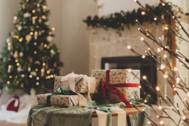 Mutlu Noeller ve mutlu tatiller! Süslü Noel hediyeleri, arka planda süslemeli Noel ağacının süsleri, kırsal şömineler ve şenlikli ışıklarla süslenmiş kurdeleli.
