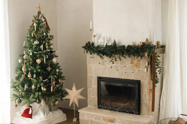 Stylish decorated fireplace mantel with christmas branches, decorations and bells on background of modern christmas tree with lights. Rustic christmas living room in modern farmhouse