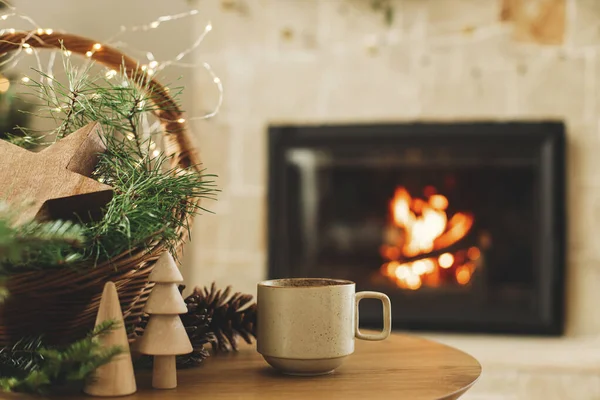 Winter cozy living room in farmhouse. Stylish cup of warm tea, basket with fir branches, wooden trees and star, pine cones against burning fireplace. Modern christmas rustic eco friendly decor
