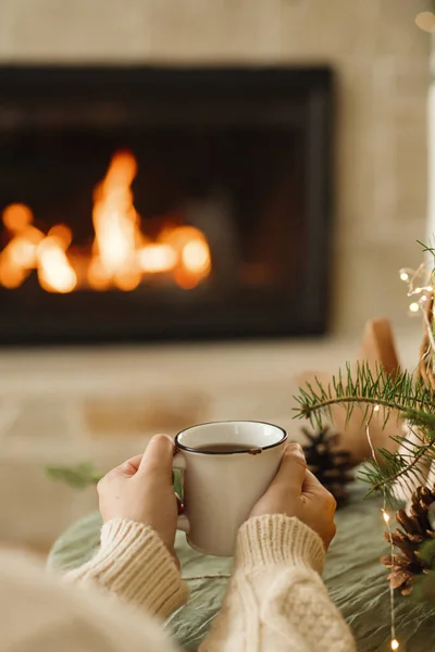 Hands in cozy sweater holding warm cup of warm tea on table with fir branches, wooden trees and and lights against burning fireplace. Atmospheric cozy christmas time. Winter hygge
