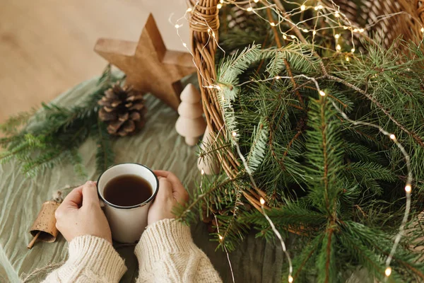 Hands in cozy sweater holding warm cup of warm tea on table with fir branches in rustic basket, wooden trees and star, pine cones and lights. Atmospheric cozy christmas time. Winter hygge