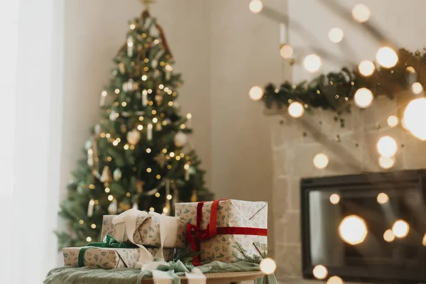 ヴィンテージバブル 素朴な暖炉 お祝いのライトが付いている現代装飾されたクリスマス ツリーの背景のテーブルのリボンが付いているスタイリッシュな包まれたクリスマスのギフト メリークリスマスとハッピーホリデー — ストック写真
