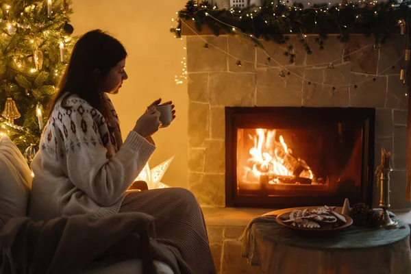 Woman in cozy sweater holding cup of tea and warming up at burning fireplace on background of stylish christmas tree with lights in evening festive room. Winter holidays hygge. Christmas eve