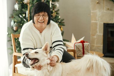 Güzel, olgun bir kadın şık bir Noel ağacı ve şenlikli şöminenin arka planında sevimli beyaz köpekle oynuyor. Mutlu yetişkin kadın evcil hayvanla sıcak kış tatilinin tadını çıkarıyor.