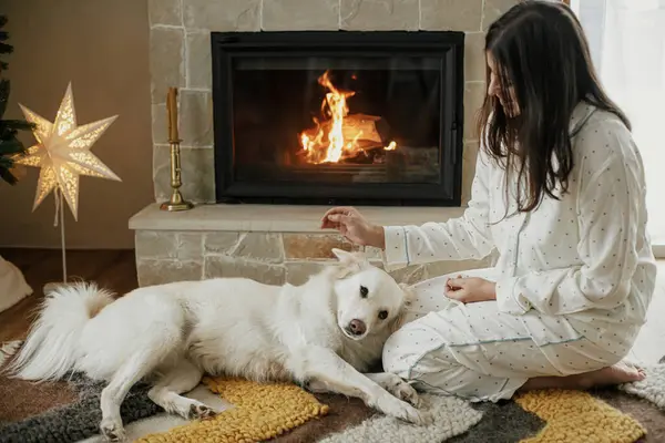 キレイなクリスマスの朝 お祝いの装飾されたリビングルームで暖かい暖炉でかわいい白い犬とリラックスして遊ぶスタイリッシュなパジャマの美しい女性 メリークリスマス ペットとの冬の休日 — ストック写真