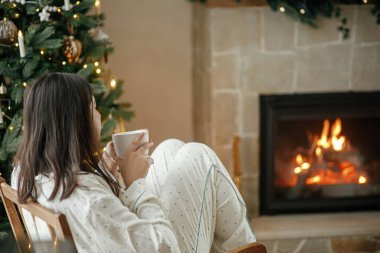 Sıcak bir Noel sabahı. Şık pijamalı güzel bir kadın şöminede sıcak bir fincan çayla rahatlıyor ve şenlikli oturma odasında süslü bir ağaç süslüyor. Mutlu Noeller! Kış tatili karışımı.