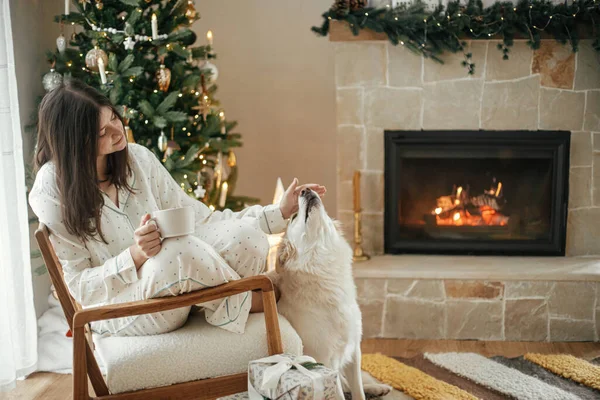 かわいい犬と居心地の良い暖かい暖炉でリラックスしてスタイリッシュなパジャマで美しい女性は お祝いの装飾されたリビングルームでプレゼントを楽しんでいます メリークリスマス ウィンター — ストック写真