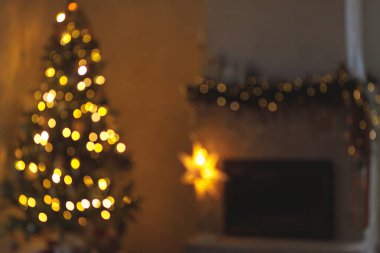 Noel arkaplanı altın bokeh. Atmosferik Noel arifesi şöminede. Aydınlatılmış yılbaşı ağacı ve akşam İskandinav odasındaki ışıklarla süslenmiş şöminenin bulanık görüntüsü