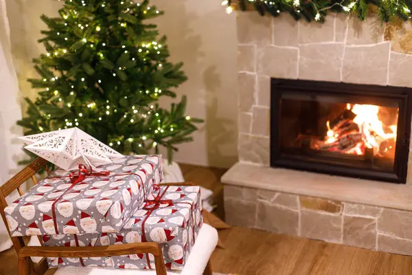 Stylish wrapped christmas gifts on arm chair against christmas tree with festive lights and cozy burning fireplace. Christmas eve, holiday time. Atmospheric living room with xmas presents