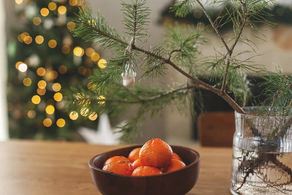 Ahşap kasede taze mandalinalar ve ladin dalı, İskandinav odasında altın ışıklarla süslenmiş şık Noel ağacına karşı. Festival evinde atmosferik kış tatili