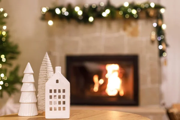 Stylish white house and tree decor in festive room, christmas eve time. Christmas modern decoration on table against burning fireplace. Atmospheric christmas time.