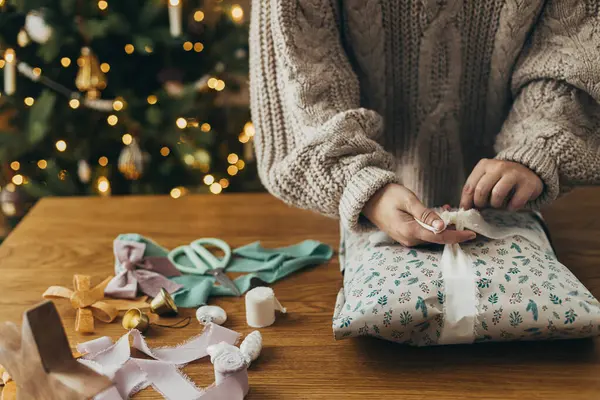 Wrapping christmas gifts. Hands in cozy sweater wrapping stylish present in festive wrapping paper with ribbons, vintage ornaments, bows on wooden table. Atmospheric winter holidays