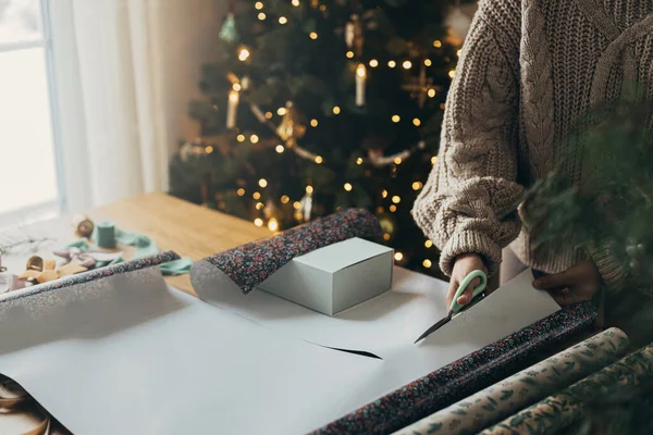 Wrapping christmas gifts. Hands in cozy sweater cutting stylish festive wrapping paper with ribbons, vintage ornaments, bows on wooden table. Atmospheric winter holidays