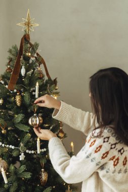 Noel ağacını şık takılarla süslemek. Ağaç dalına asılmış, rahat süveterli bir kadın. Atmosferik kış tatili geleneği, aile zamanı