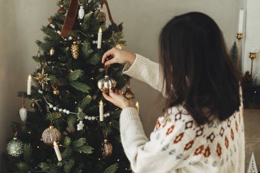 Noel ağacını şık takılarla süslemek. Ağaç dalına asılmış, rahat süveterli bir kadın. Atmosferik kış tatili geleneği, aile zamanı