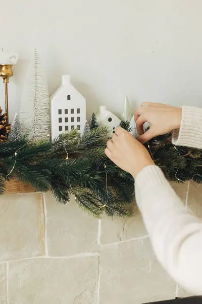 Decorating christmas fireplace with lights, fir branches, white little houses decoration. Hands in cozy sweater hanging decoration on mantel in modern farmhouse. Atmospheric winter holiday