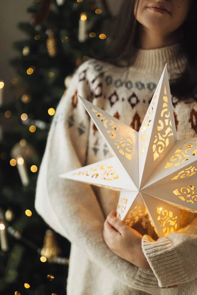 Stylish glowing big christmas star in hands of on background of decorated christmas tree with vintage baubles and lights. Merry Christmas! Atmospheric magic winter time