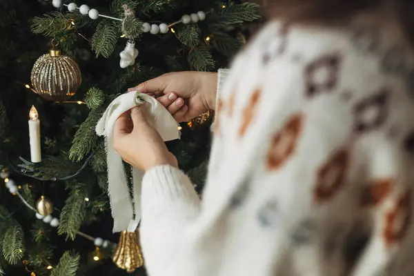 スタイリッシュなリボンでクリスマスツリーを飾ります 木の枝に近代的な白い装飾を掛ける居心地の良いセーターの手が近づいています 大気の冬の休日の伝統 家族の時間 — ストック写真