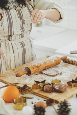 Elleri ahşap tahta tahtada zencefilli ekmek hamuru, merdane, altın metal kesiciler, yemek baharatları, modern beyaz mutfakta şenlikli süslemeler. Kadın Noel kurabiyesi yapıyor.