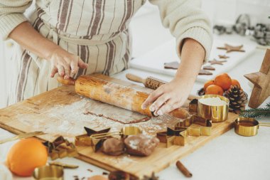 Elleri ahşap tahta tahtada zencefilli ekmek hamuru, merdane, altın metal kesiciler, yemek baharatları, modern beyaz mutfakta şenlikli süslemeler. Kadın Noel kurabiyesi yapıyor.