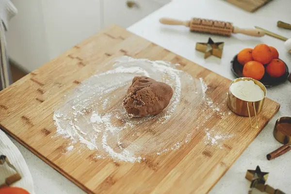 Gingerbread dough with flour on wooden board, rolling pin, golden metal cutters, cooking spices and festive decorations on countertop in modern white kitchen. Making christmas gingerbread cookies