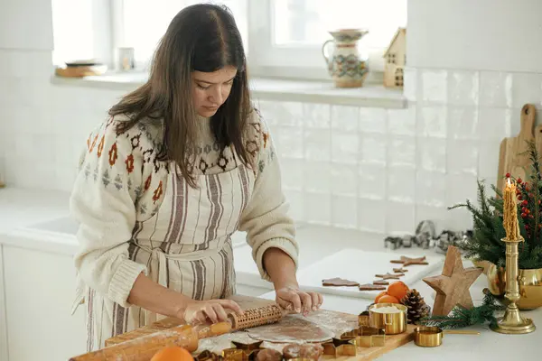 Woman making christmas gingerbread cookies in modern white kitchen. Hand kneading gingerbread dough on wooden board with flour, rolling pin, golden metal cutters, cooking spices on countertop