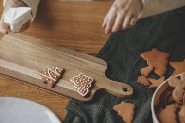 Kırsal ahşap masa üzerindeki kremalı zencefilli kurabiyeleri yakından süslemek. Atmosferik Noel gelenekleri. Elleri şekerli kurabiyelerle fırında pişmiş Noel kurabiyeleri süslüyordu. Aile zamanı.