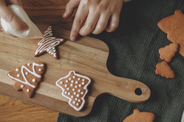Eller, Noel kurabiyelerini kırsal ahşap masa manzaralı kremayla süslüyor. Atmosferik Noel gelenekleri. Kurabiyeleri şekerli süslemek. Aile zamanı.