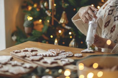 Elleri zencefilli kurabiyeleri kırsal ahşap masa üzerinde kremayla süslüyor. Noel altın ışıklarının arka planında. Atmosferik Noel gelenekleri. Kurabiyeleri şekerli süslemek