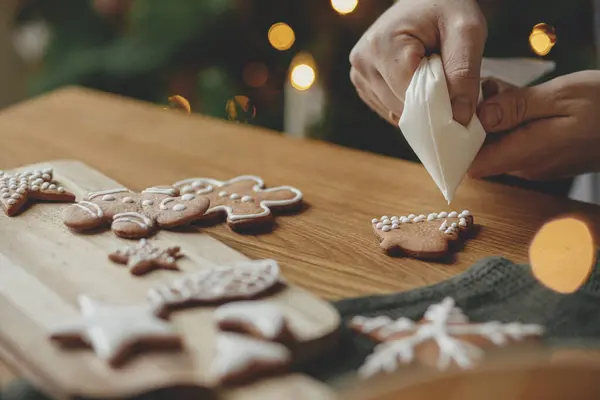 Decorating gingerbread cookies with icing on rustic wooden table at christmas tree golden lights. Atmospheric Christmas holiday traditions. Man decorating cookies with sugar frosting