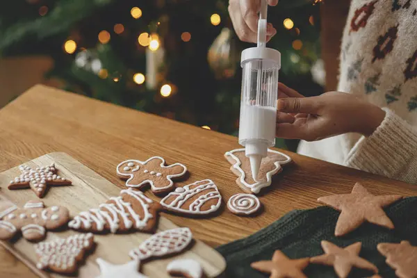 Decorating gingerbread cookies with icing on rustic wooden table at christmas tree golden lights. Atmospheric Christmas holiday traditions. Woman decorating cookies with sugar frosting