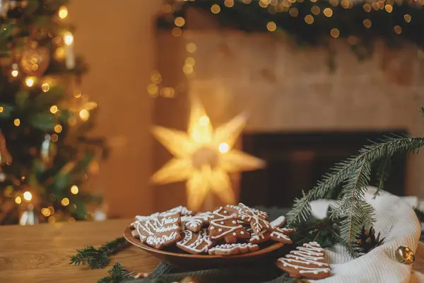 メリークリスマス ジンジャーブレッドクッキー 木のテーブルのプレートでアイシングとクリスマスゴールデンライトに対する枝やお祝いの装飾 大気のクリスマスイブ 家族の時間 — ストック写真