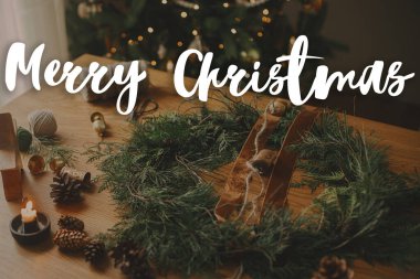 Sedir dallı, kurdeleli, masada klasik çanlar olan şık Noel kırsal çelengi üzerine Mutlu Noeller metni. İyi tatiller! Sezonun tebrik kartı. El yazısı işareti