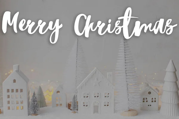 圣诞快乐的文字在时尚的小白色房子和树木 雪白的圣诞小村庄与金色的灯光 节日快乐 季节的贺卡 手写签名 — 图库照片