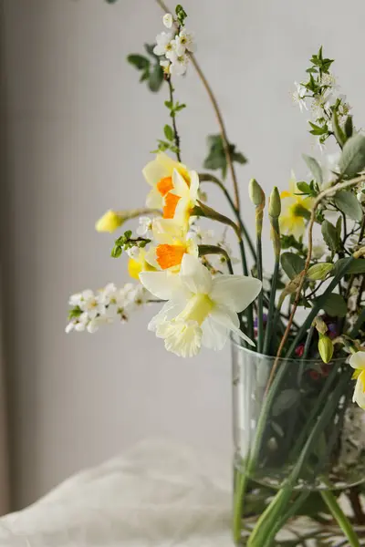 Stylish spring flowers bouquet on rustic table in rural room. Beautiful daffodils, cherry bloom and greenery composition in glass vase. Easter modern simple decor